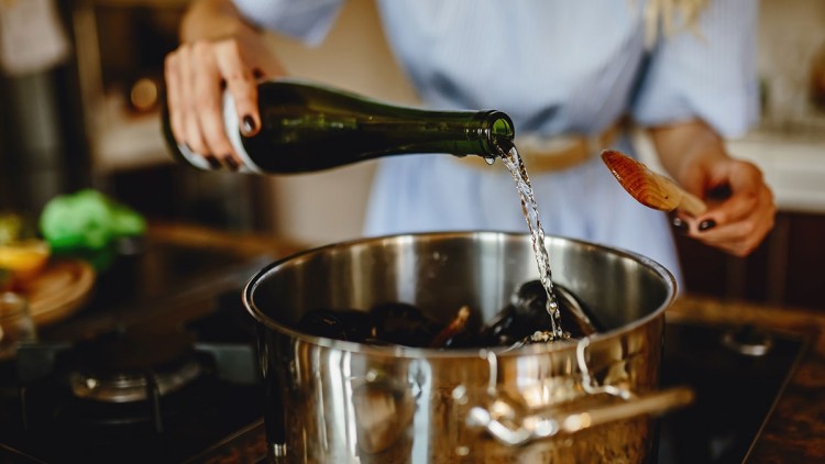 Eine Hand gießt Flüssigkeit aus einer grünen Flasche in einen Metalltopf mit Muscheln, daneben ein Holzlöffel in der anderen Hand.