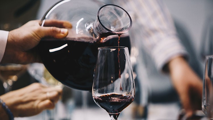 Roter Wein wird aus einer bauchigen Glaskaraffe in ein halbvolles Weinglas eingeschenkt, im Hintergrund unscharfe Personen.
