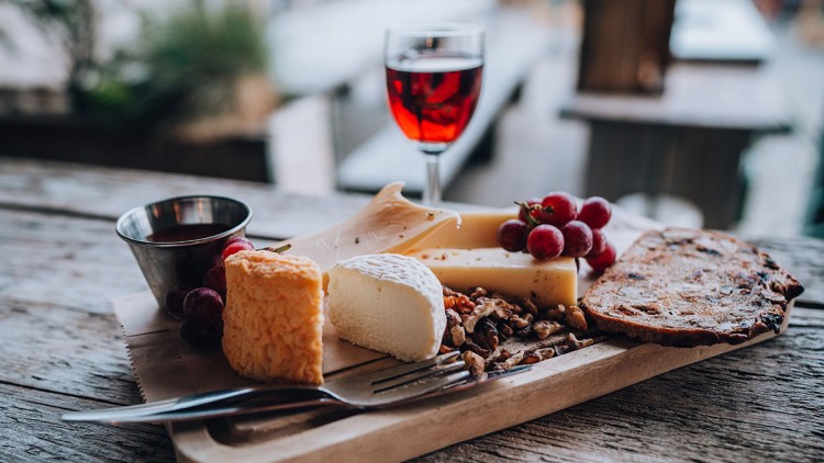 Verschiedene Käsesorten, Trauben, Nüsse, Honig und eine Scheibe Brot auf einem Holzbrett mit Glas Rotwein im Hintergrund.