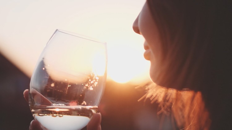 Ein halb gefülltes Weinglas wird in der Hand gehalten, im Hintergrund scheint die Sonne warm durch die Glasscheibe.