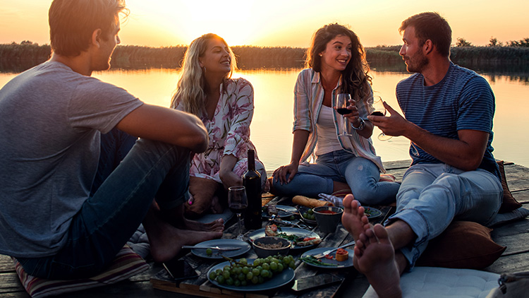 Vier Personen sitzen auf einem Holzsteg am Wasser bei Sonnenuntergang und trinken aus Gläsern mit Wein.