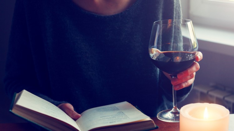 Person liest ein Buch, hält ein Glas Rotwein in der Hand, daneben brennt eine Kerze auf einem Tisch.
