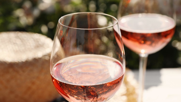Zwei gefüllte Weingläser mit roséfarbenem Getränk stehen auf einem Tisch, im Hintergrund ein strohfarbenes Accessoire.