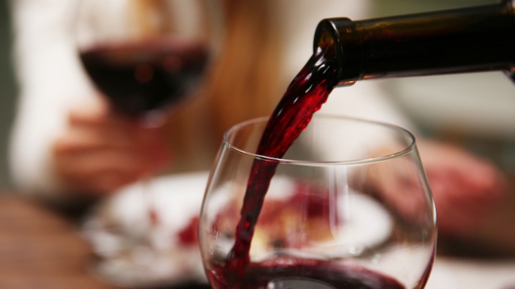 Rotwein wird aus einer Flasche in ein Weinglas eingeschenkt, im Hintergrund ist eine Person mit Weinglas unscharf zu erkennen.