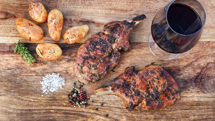 Zwei gegrillte Koteletts mit Kräutern, goldbraune Kartoffeln, Pfefferkörner, grobes Salz, Rosmarinzweig und Rotweinglas auf Holzbrett.