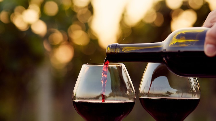 Eine Hand gießt Rotwein aus einer Flasche in eines von zwei Weingläsern, im Hintergrund unscharfe Natur und Sonnenlicht.