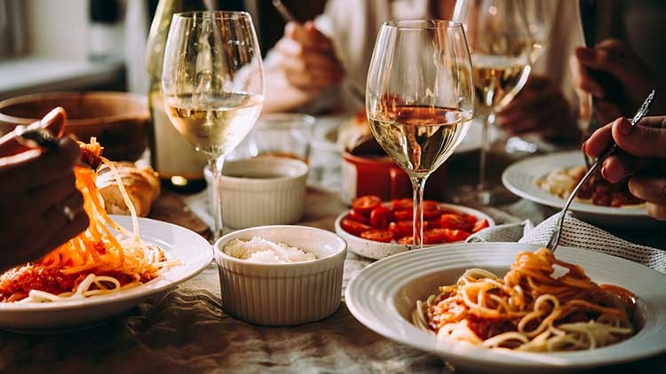 Mehrere Teller mit Spaghetti, Schälchen mit Käse und Tomaten sowie gefüllte Weingläser auf einem gedeckten Tisch.