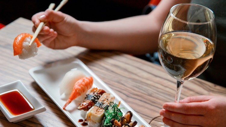 Eine Hand hält ein Glas Weißwein, die andere nimmt mit Stäbchen Sushi von einem Teller neben einer Sojaschale.