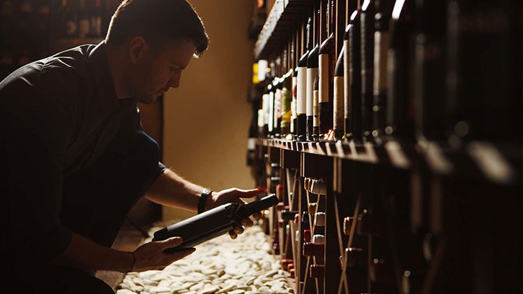 Person kniet vor Holzregal mit Weinflaschen und betrachtet eine Flasche, der Boden ist mit hellen Steinen ausgelegt.