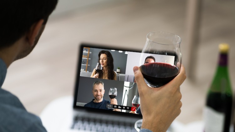 Person hält ein Glas Rotwein vor einem Laptop, auf dem eine Videokonferenz mit weiteren Personen zu sehen ist, daneben Weinflaschen.