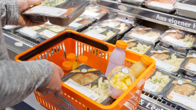 Eine Person legt eine Fertigmahlzeit in einen orangefarbenen GLOBUS-Einkaufskorb mit Getränken und Obstbecher vor einer Kühltheke.