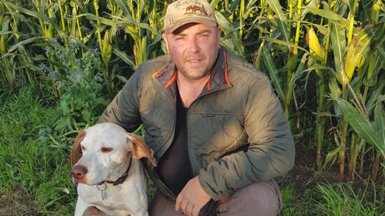 Person in grüner Jacke kniet neben einem sitzenden Hund mit weißem Fell und braunen Flecken vor einem Maisfeld.