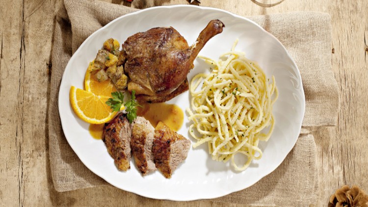Gebackene Entenkeule mit geschnittenem Entenbraten, Orangenscheibe, Soße, Knödelwürfeln und Spätzle auf weißem Teller.