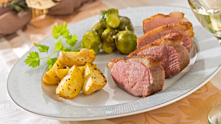 Ein weißer Teller mit knusprigem Fleisch, gerösteten Kartoffelstücken, Rosenkohl und einem Zweig frischer Petersilie.