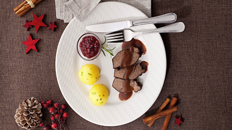 Weißer Teller mit Fleisch in brauner Sauce, zwei Klößen, Preiselbeersauce im Glas, Besteck und roten Sternen auf braunem Stoff.