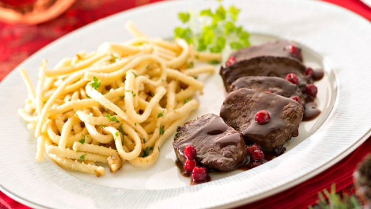 Ein weißer Teller mit Spätzle, verziert mit Petersilie, und drei Fleischstücken in dunkler Sauce mit roten Beeren.
