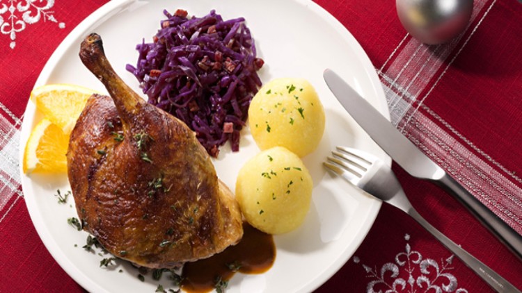 Gedeckter Teller mit Entenkeule, Bratensauce, zwei Kartoffelklößen, Rotkohl und Orangenscheiben auf rotem Tischtuch.