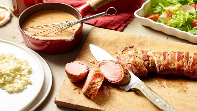 Ein Holzbrett mit aufgeschnittenem Fleisch, daneben ein Messer, eine rote Soßenkasserolle, Risotto und ein Salat in weißer Schale.