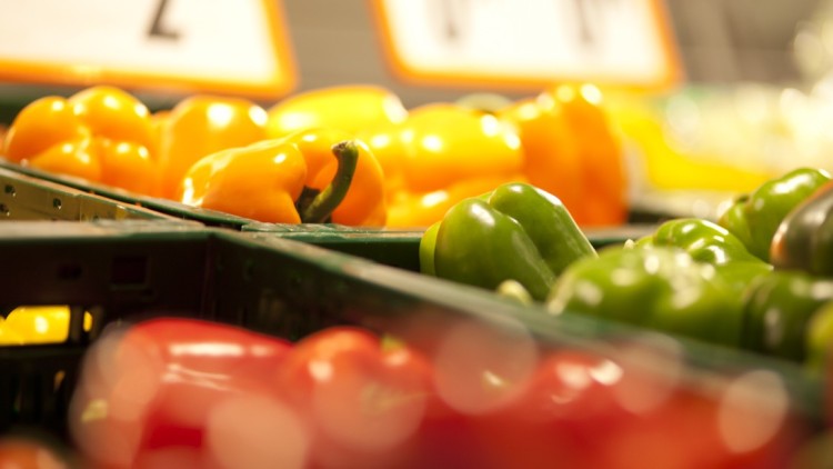 Frische Paprika in den Farben Gelb, Grün und Rot liegen nebeneinander in grünen Gemüsekisten in einer Auslage.