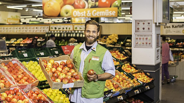 Mitarbeiter im grünen GLOBUS-Westen hält eine Kiste mit roten Äpfeln vor Regalen in der Obst- und Gemüseabteilung.