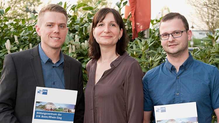 Drei Personen stehen vor grüner Hecke, zwei halten eine Mappe mit der Aufschrift „Bildungszentrum der IHK Bonn/Rhein-Sieg“.