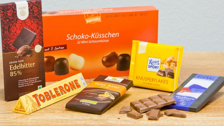 Verschiedene Schokoladenprodukte, darunter Tafeln, Schaumküsse und ein gelber Toblerone-Riegel, auf einem Holztisch.