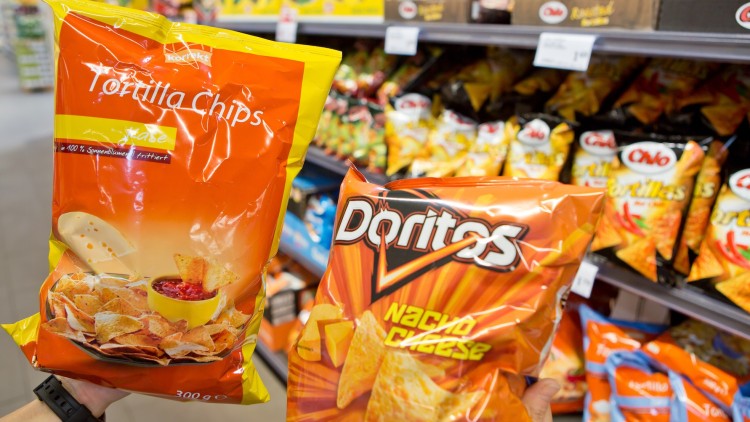 Zwei Hände halten je eine Tüte Tortilla Chips der Marken „korrekt“ und „Doritos“ vor einem Supermarktregal.