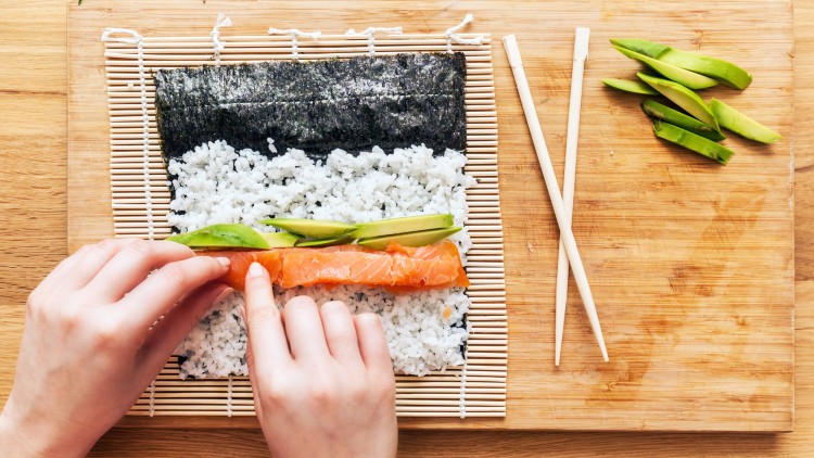 Hände rollen auf einer Bambusmatte ein Sushi mit Noriblatt, Reis, Lachs und Avocadoscheiben, daneben Holzstäbchen und Avocado.