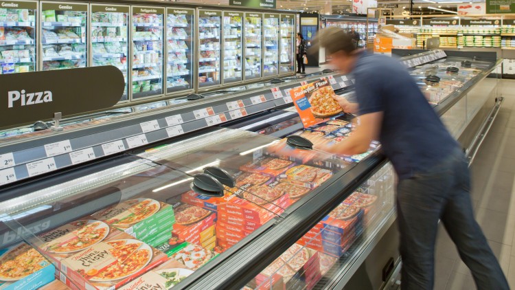 Person greift in Kühltheke mit Tiefkühlpizzen, Hintergrund zeigt weitere Kühlregale mit Gemüse und Fertiggerichten.