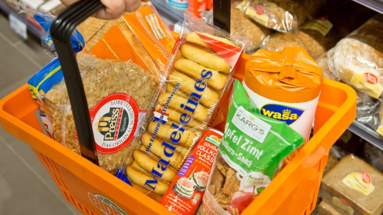 Oranger Einkaufskorb mit Lebensmitteln wie Brot, Madeleines, Keks-Snacks und Wasa-Produkten, gehalten von einer Hand.