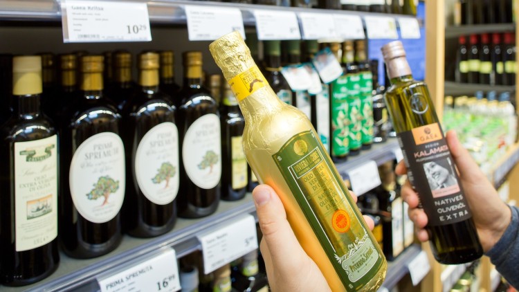 Zwei Hände halten Flaschen mit Olivenöl vor einem Supermarktregal mit verschiedenen Olivenölflaschen und Preisschildern.