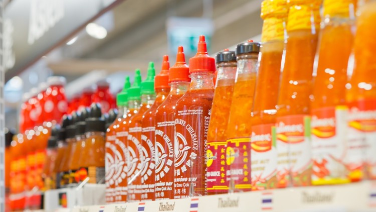 Regal mit verschiedenen Chilisaucen in Glasflaschen, Beschriftung „Thailand“ und kleine Landesflaggen auf den Preisschildern.