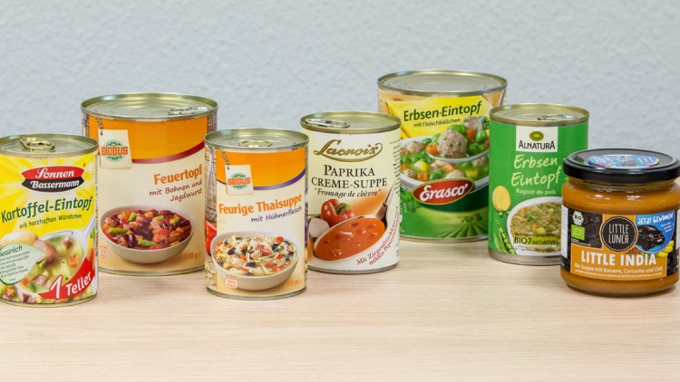 Sieben Suppen- und Eintopfprodukte in Dosen und Gläsern, darunter Marken wie GLOBUS, Erasco und Alnatura, auf Holzuntergrund.