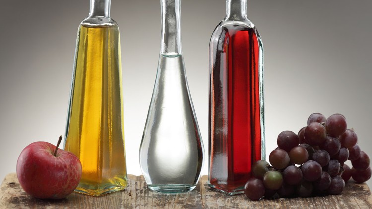 Drei Glasflaschen mit goldenem, klarem und rotem Inhalt stehen auf Holz neben einem roten Apfel und einer roten Traube.