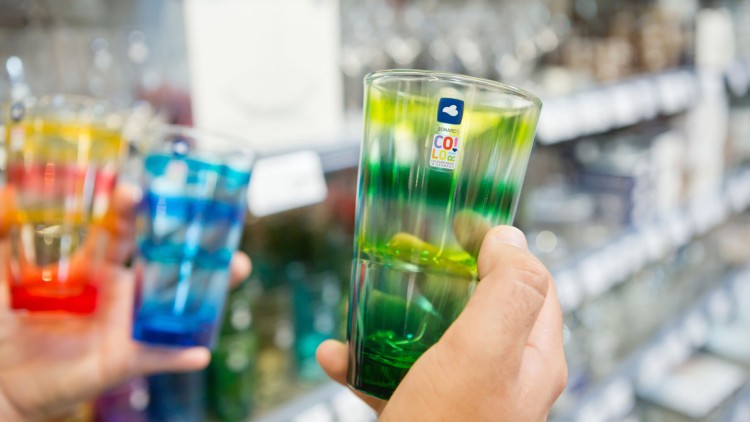 Eine Hand hält ein grün-gelbes Trinkglas mit „Leonardo COLO“-Aufkleber, dahinter weitere bunte Gläser und Regale.
