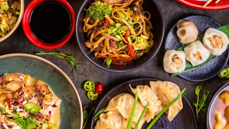 Verschiedene asiatische Gerichte wie Dumplings, gebratene Nudeln, Reis, Dim Sum und Suppe mit frischen Beilagen.