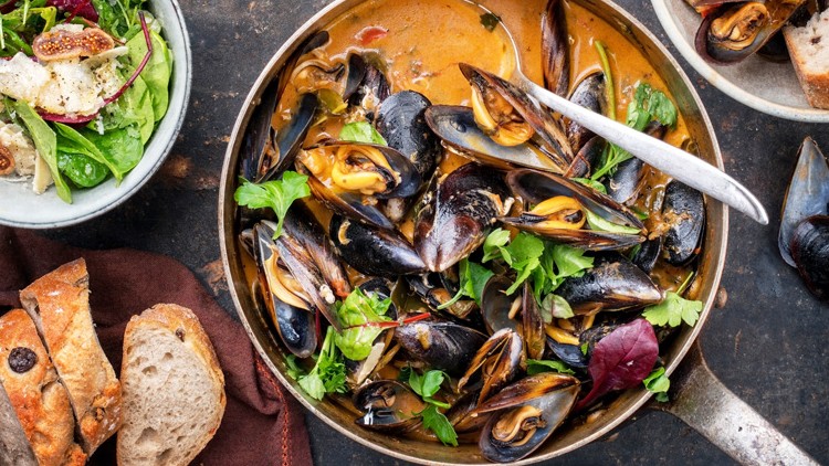 Pfanne mit Muscheln in heller Sauce und Petersilie, dazu Salat mit Feigen und Brot mit Oliven auf dunklem Untergrund.