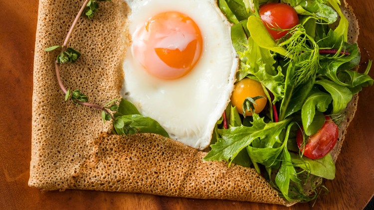 Brauner Crêpe auf Holzplatte, gefüllt mit Spiegelei, grünem Salat, gelben und roten Cherrytomaten sowie frischen Kräutern.