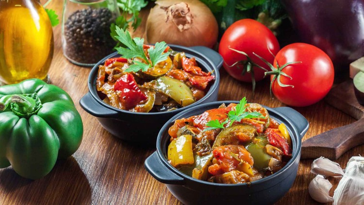 Zwei schwarze Schalen mit Gemüseeintopf stehen auf einem Holztisch, umgeben von Paprika, Tomaten, Aubergine, Zwiebel und Kräutern.