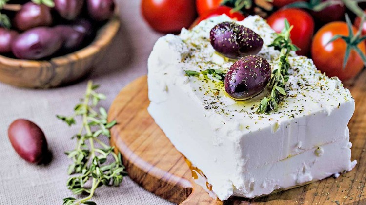 Ein Stück weißer Feta mit Kräutern, Olivenöl und zwei dunklen Oliven auf einem Holzbrett, daneben Zweige und Kirschtomaten.