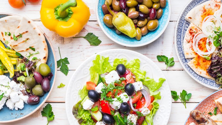 Verschiedene mediterrane Gerichte mit Salat, Oliven, Feta, Gemüse, Fladenbrot und gegrillten Speisen auf weißem Tisch.
