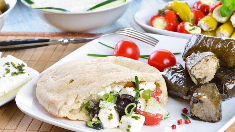 Teller mit gefülltem Fladenbrot, Weinblättern, Käsewürfeln, Oliven, Tomaten und Kräutern, im Hintergrund weitere mediterrane Speisen.