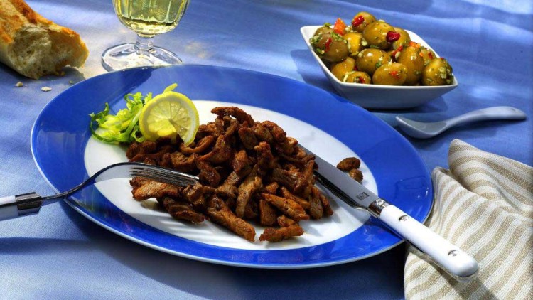 Gebratene Fleischstreifen mit Zitronenscheibe und grünem Salat auf blau-weißem Teller, daneben Olivenschale und Glas Weißwein.