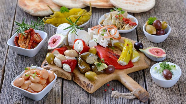 Eine Holzplatte mit Antipasti wie gefüllten Paprikaschoten, Oliven, Käse, Feigen und Schälchen mit weiteren Beilagen.