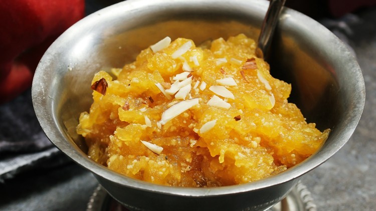 Silberne Schale mit gelbem Dessert aus geriebenem Obst, garniert mit Mandelstücken, daneben ein Löffel und ein roter Apfel.