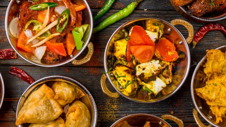 Verschiedene indische Gerichte wie Currys, Paneer, Samosas und Pakoras in Metallschalen mit Gewürzen wie Chilischoten und Fenchelsamen.