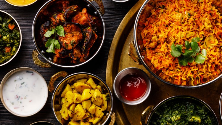 Verschiedene indische Gerichte wie Curry, Reis, Gemüse, Fladenbrot und Saucen in Schalen und Tellern auf dunklem Untergrund.