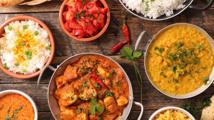 Verschiedene indische Gerichte in Schüsseln, darunter Curry, Reis, Linsen, Tomaten und Fladenbrote auf einem Holztisch.