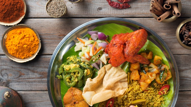 Ein Edelstahl-Teller mit Gewürzreis, Gemüse, frittiertem Teig, Curry, Salat und einem roten Hähnchenschenkel auf Holzuntergrund.