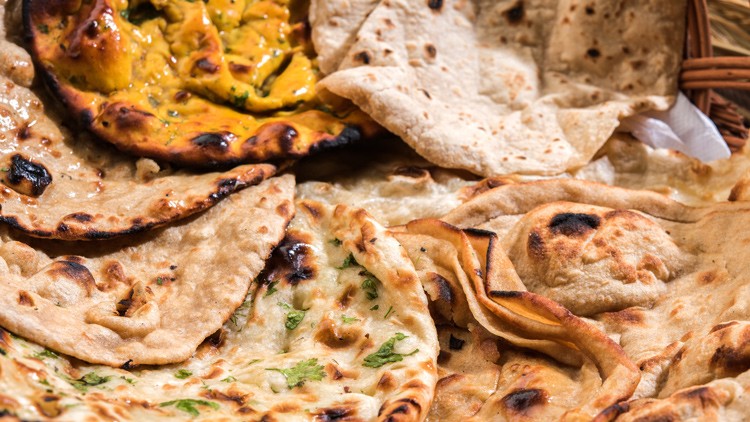 Verschiedene Sorten indisches Fladenbrot wie Naan und Paratha liegen in einem Korb auf einem Holztisch.